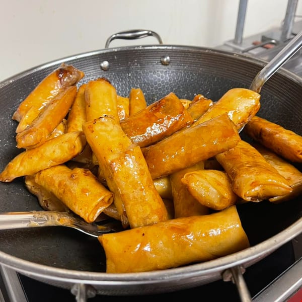 A large pan filled with golden-brown, glazed Lumpia House spring rolls, with a serving spoon.