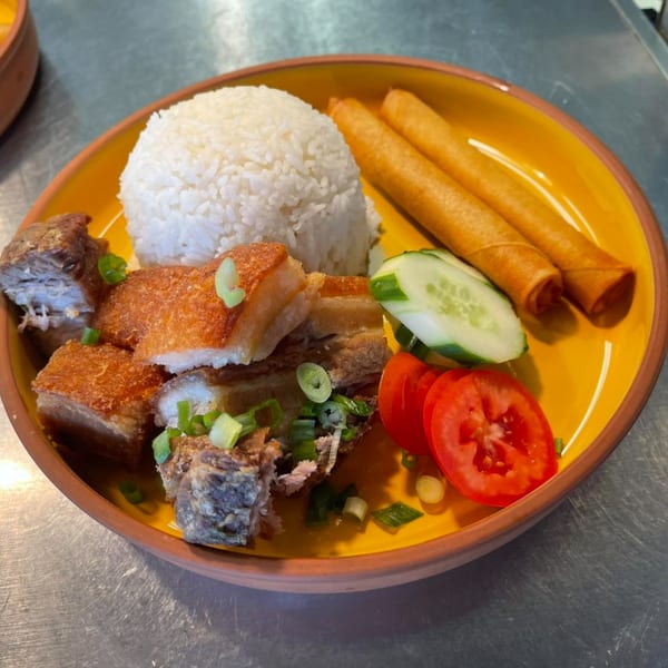 Crispy pork belly, white rice, two lumpia, sliced cucumber, and tomatoes on a yellow plate.