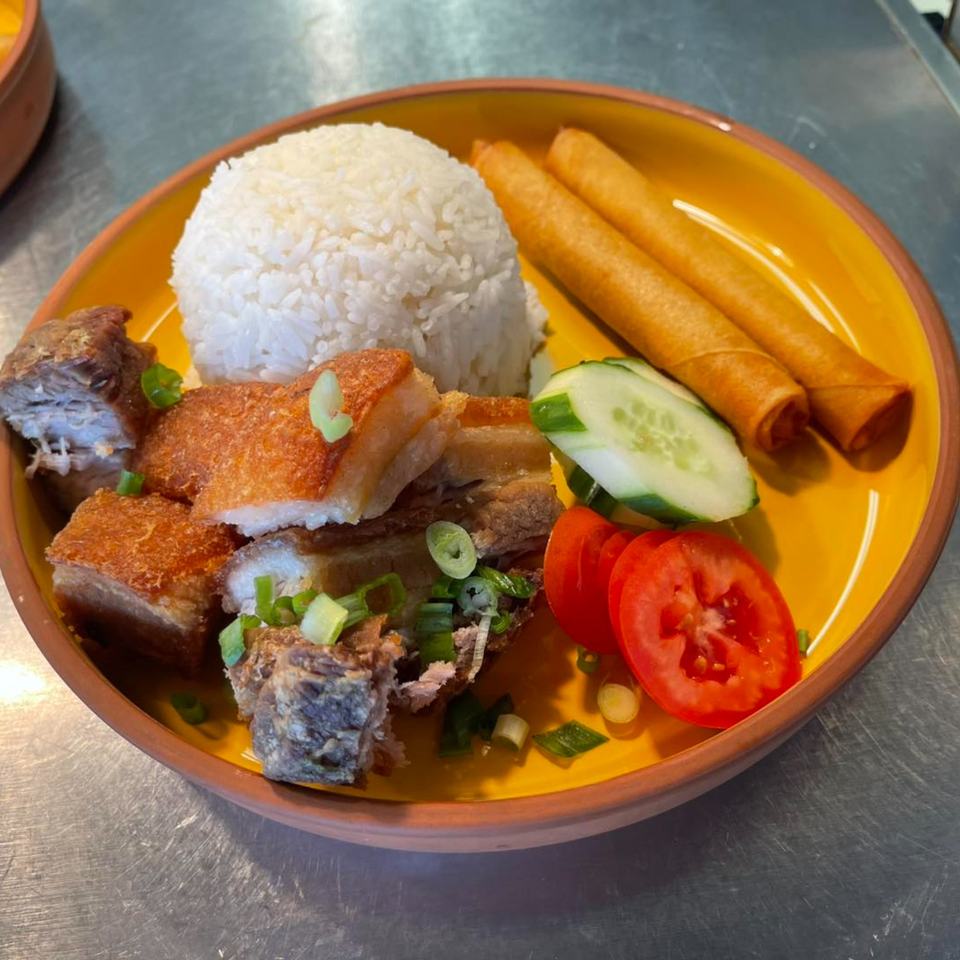 Crispy pork belly, white rice, two lumpia, sliced cucumber, and tomatoes on a yellow plate.