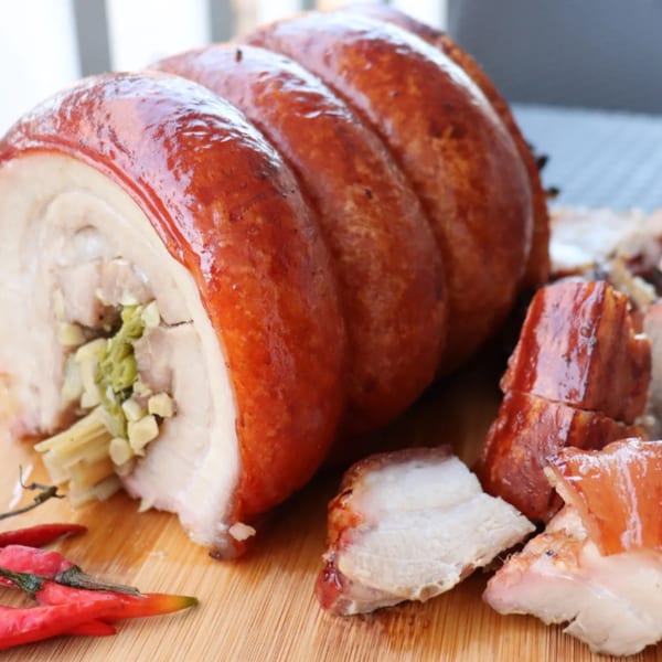 Crispy rolled pork belly lechon with herbs, sliced and served on a wooden board with red chilies.