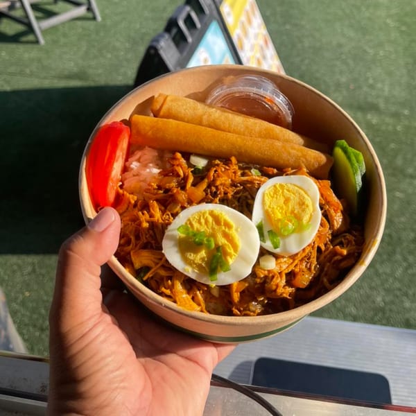Hand holding a bowl of Asian food with lumpia, chicken adobo, rice, and hard-boiled eggs.