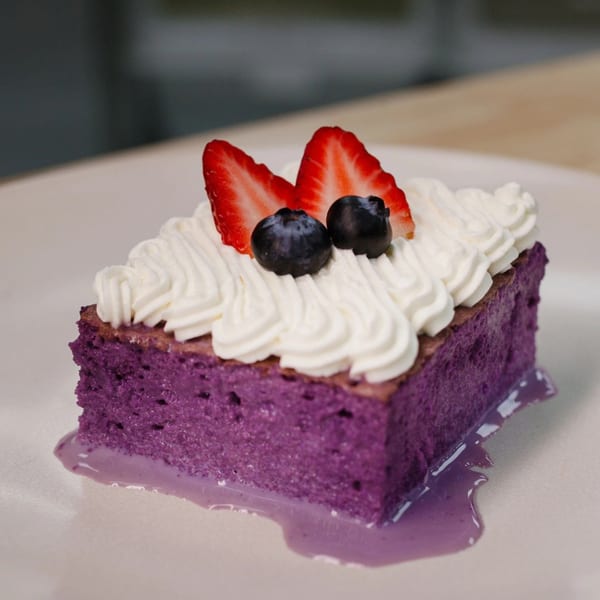 A slice of purple ube cake with whipped cream, strawberries, and blueberries on a white plate.